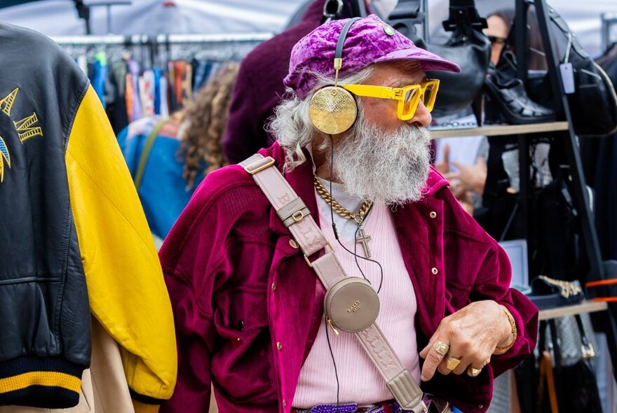 A man with a long grey beard wearing over-the-ear headphones and bright yellow glasses in an outdoor market setting. 