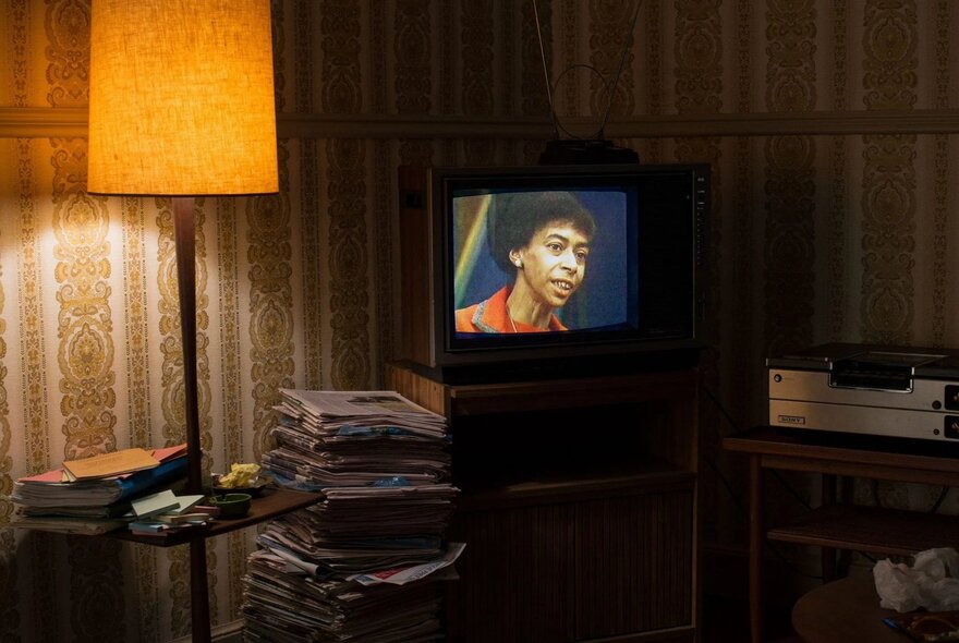 A still from the film, Recorder: The Marion Stokes Project, that depicts a room with a tube TV on in the corner, a floor lamp, wallpaper on the walls, and newspapers piled up on the floor. 