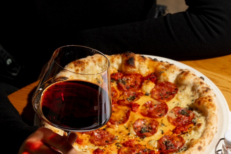 A traditional-style Italian pizza with salami and melted cheese, a glass of red wine in front.