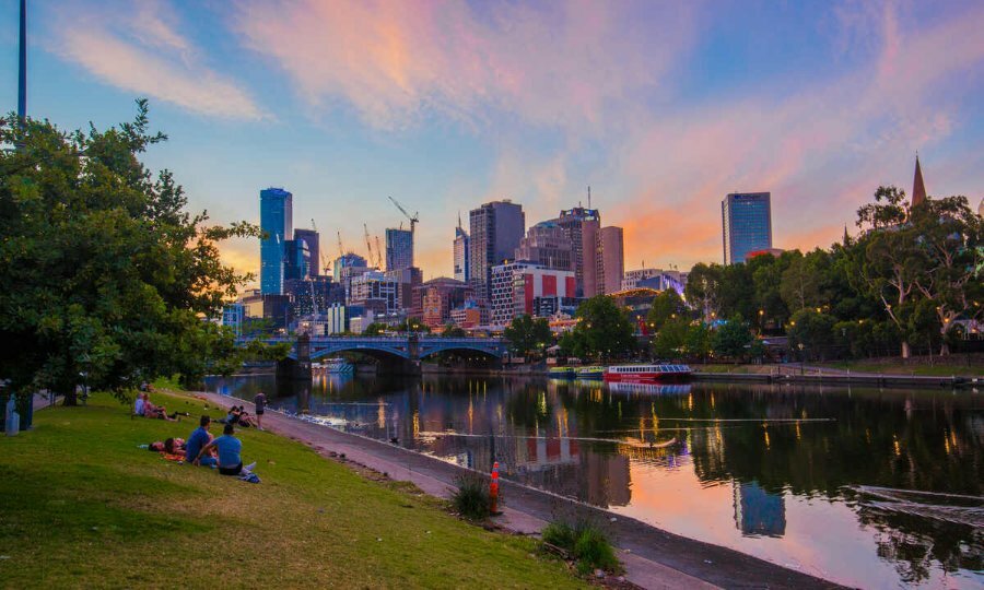 Wide shot of people sitting on grass hill watching the sunset over city skyline.