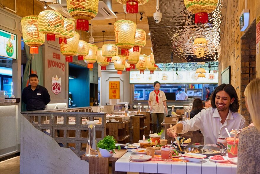 Two friends are eating hotpot in a Chinese restaurant. There are glowing lanterns hanging from the ceiling. 