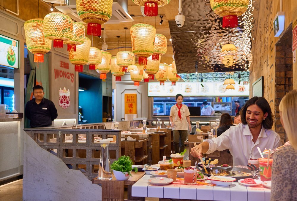 Two friends are eating hotpot in a Chinese restaurant. There are glowing lanterns hanging from the ceiling. 