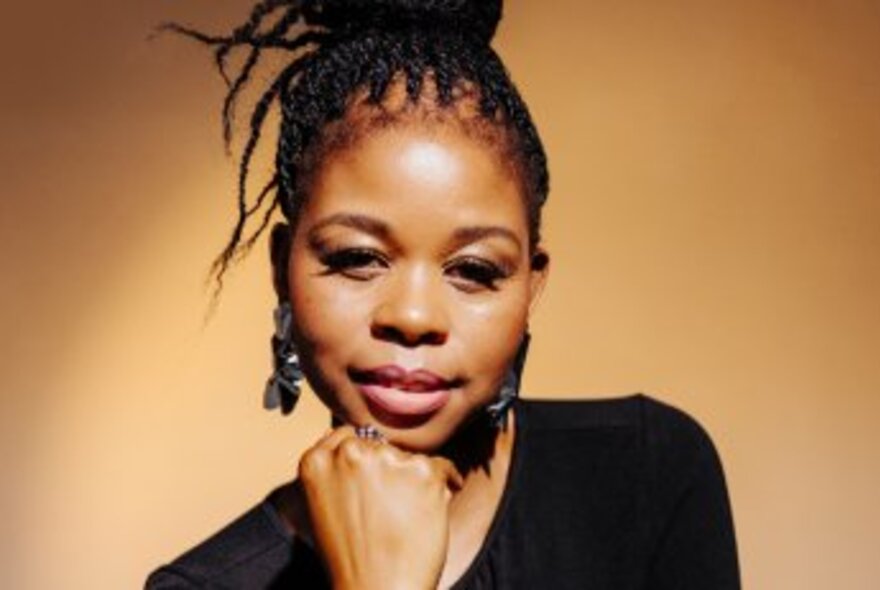 A close-up portrait of Dorcas Maphakela, a South African writer. She has her braided hair in a bun, is wearing large dark earrings and a black top, and is resting her hand on her chin.