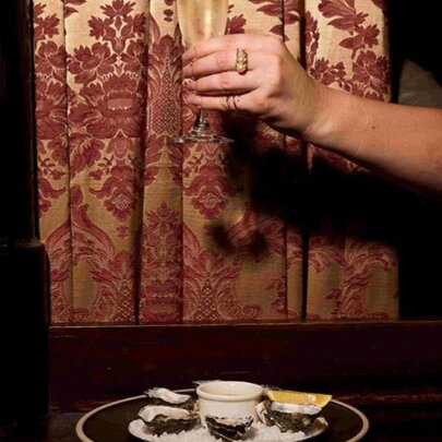 Hand holding a glass of champagne against a patterned curtain with platter of oysters.