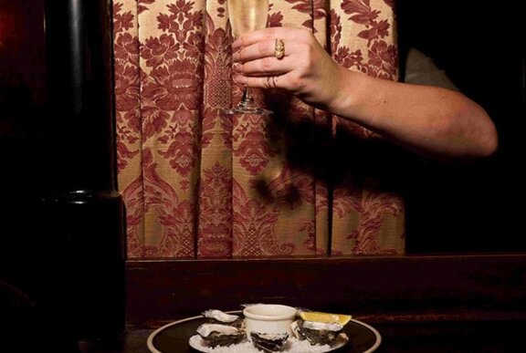 Hand holding a glass of champagne against a patterned curtain with platter of oysters.