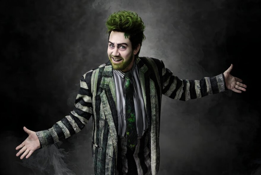 Beetlejuice actor in costume with green hair and striped grey suit, smiling with arms outstretched.