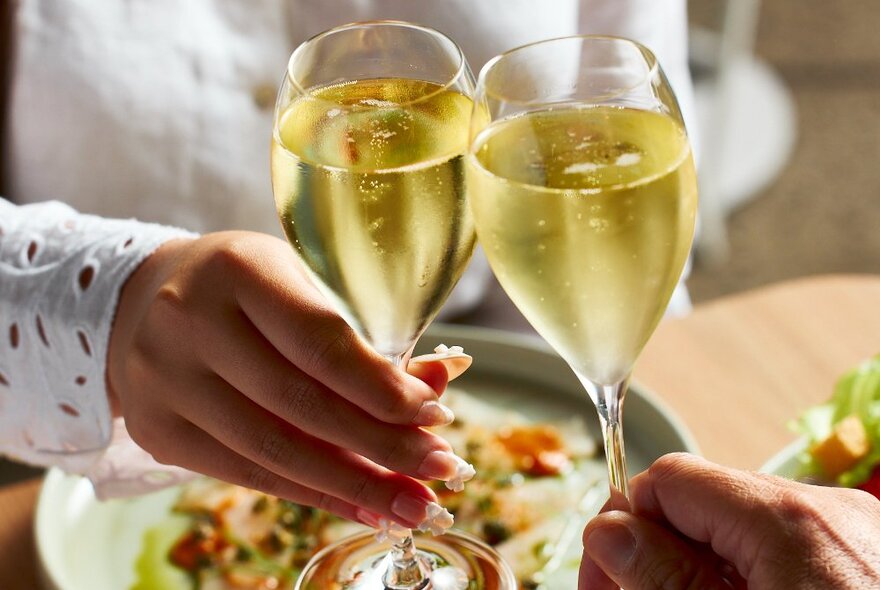 Two hands clinking glasses of sparkling wine over a plate of food.