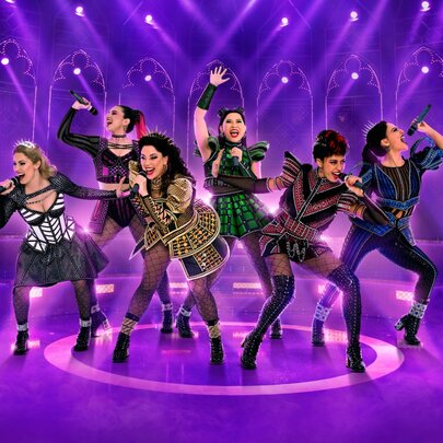 Six female performers on a purple lit stage in costumes and striking poses, all singing into microphones they hold in their hands.