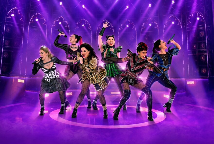 Six female performers on a purple lit stage in costumes and striking poses, all singing into microphones they hold in their hands.