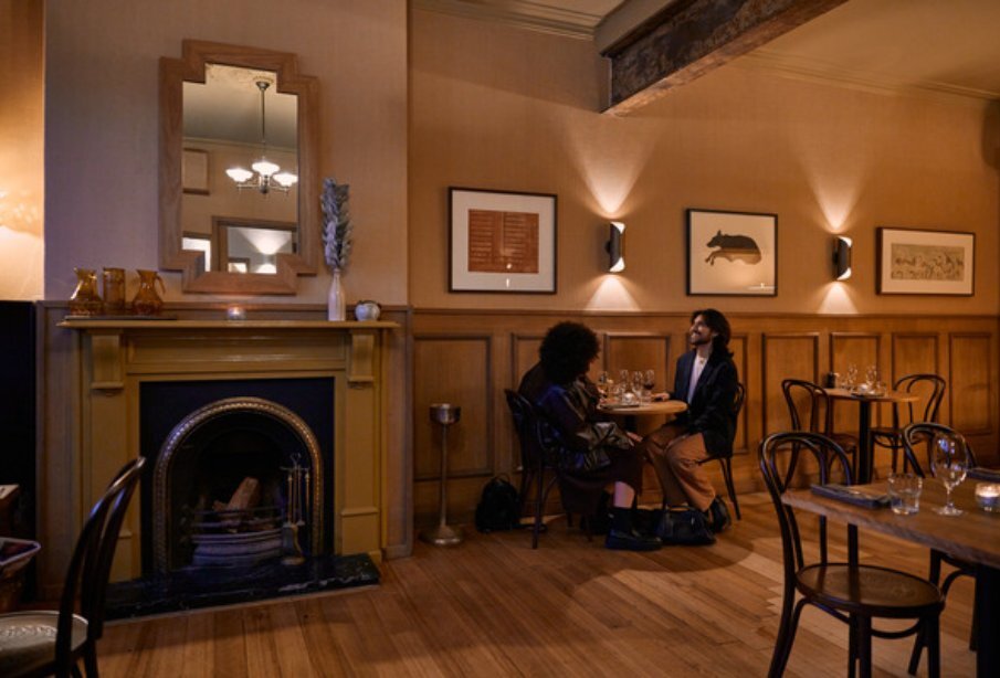 Two people seated in an art-deco style pub next to a fireplace.