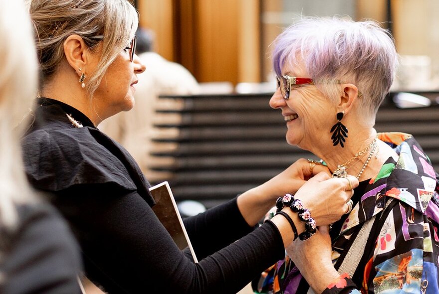 A woman fastening a necklace on another woman who has short hair, long earrings and a smile. 