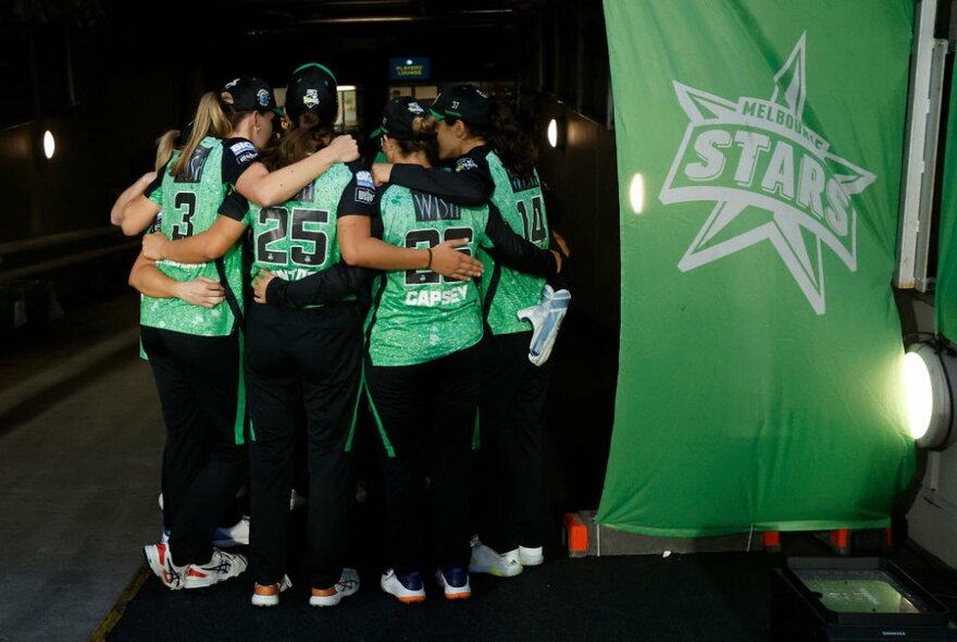 The Melbourne Stars WBBL team hugging together in a circle with a green flag next to them.