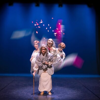 A central figure in white robes stands in a spotlight, holding a wooden cane, while three other performers in similar light-coloured costumes pose closely behind them.