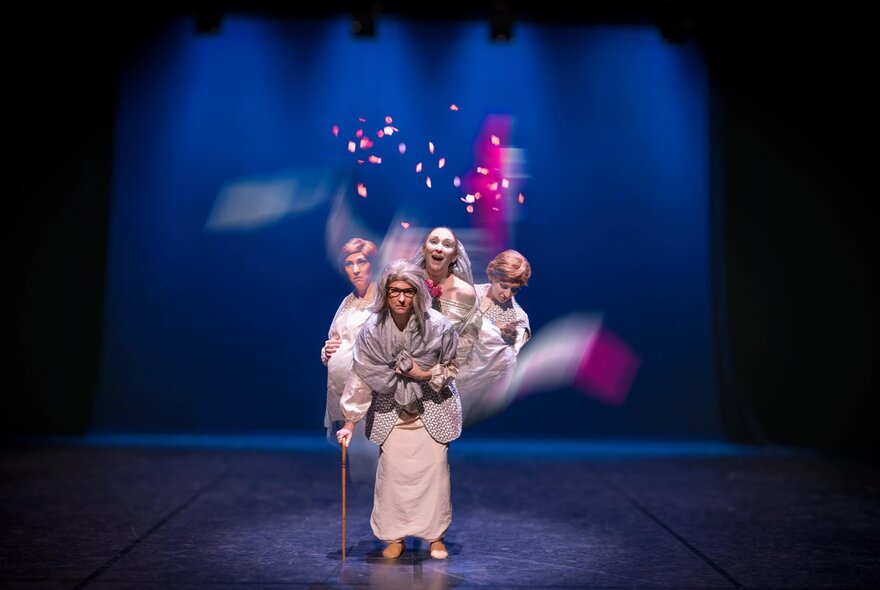 A central figure in white robes stands in a spotlight, holding a wooden cane, while three other performers in similar light-coloured costumes pose closely behind them.
