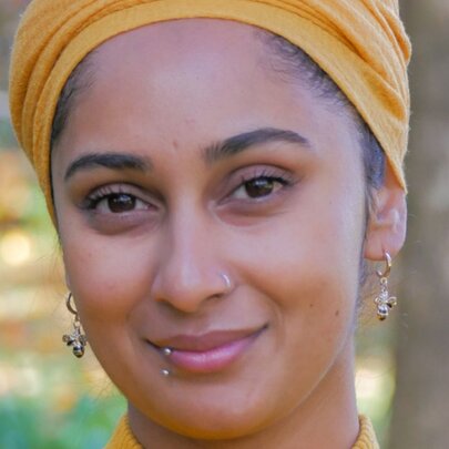 Portrait of writer Soaliha Iqbal standing outside, with earrings and a lip piercing and a yellow head scarf.
