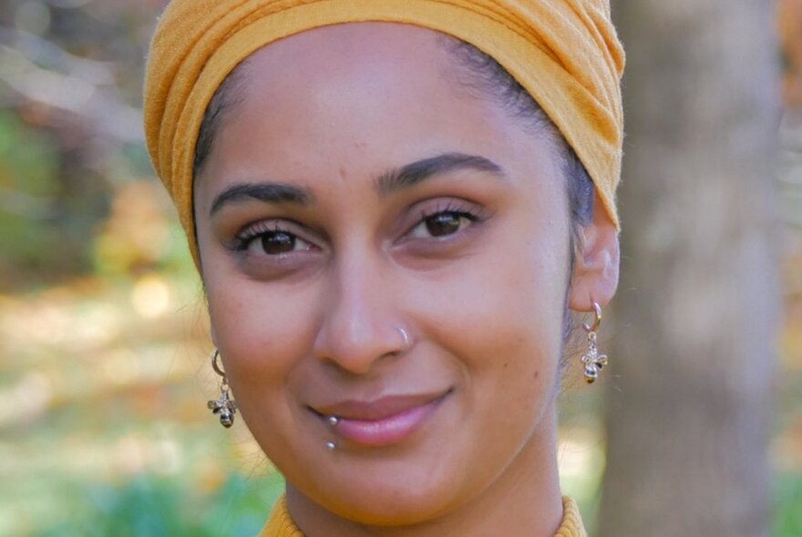 Portrait of writer Soaliha Iqbal standing outside, with earrings and a lip piercing and a yellow head scarf.