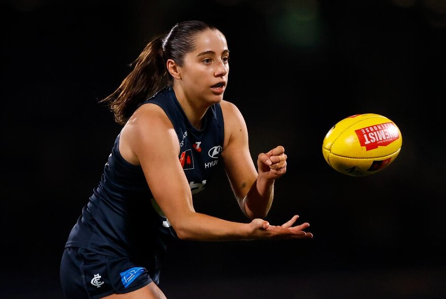 Female AFL football player handballing a yellow ball on a dark pitch.