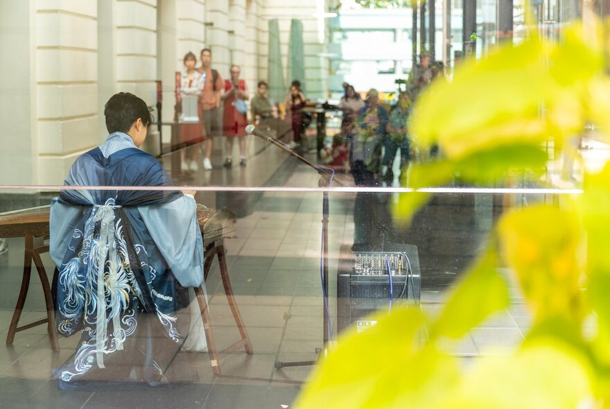 A person in a blue outfit with white tassels sits facing away, playing a string instrument in a public atrium, with other people standing in the background watching.