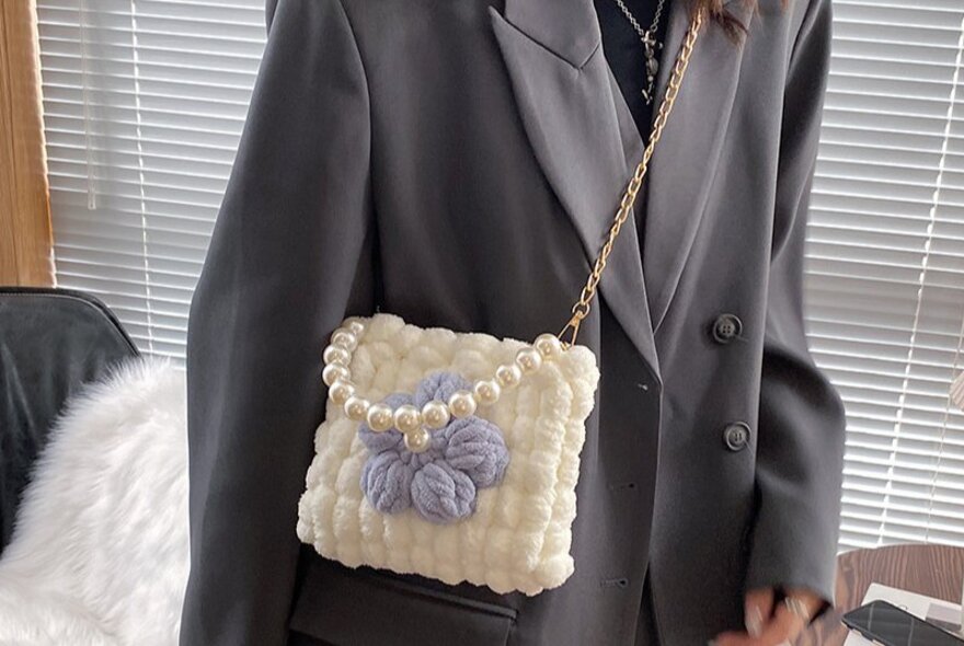 A person modelling a cream knitted shoulder bag with plush fabric and a chain strap, made in a cross-stitch workshop.