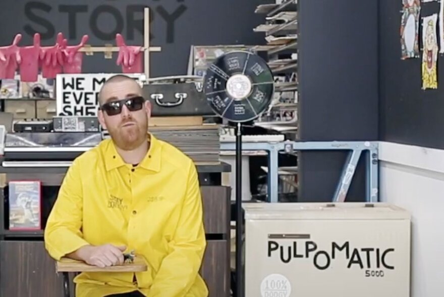 A man in a bright yellow shirt and sunglasses seated in a workshop space, a cardboard box with the words PULPOMATIC written on the sideof the box.