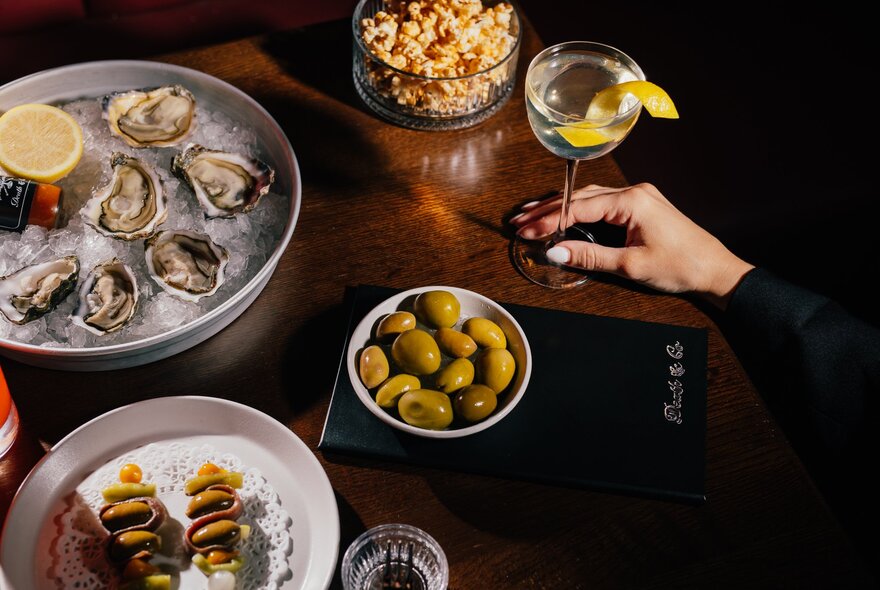 A hand holding the stem of a martini glass that rests on a small table, with a plate of oysters, a bowl of olives and other nibbles nearby.