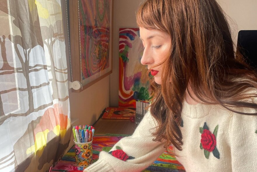 A young adult at a desk near a window, with a jar of coloured pens on the table.