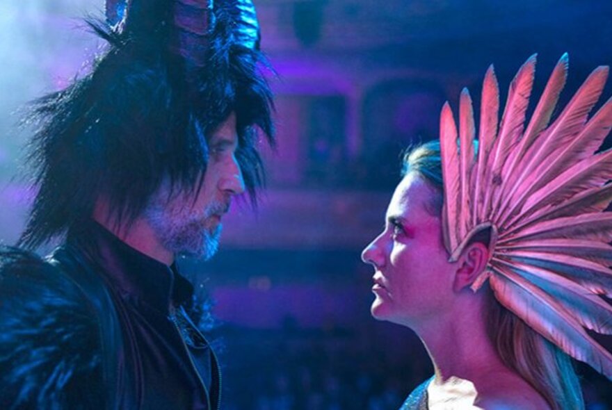 Man and woman wearing featherlike headresses, staring at one another.