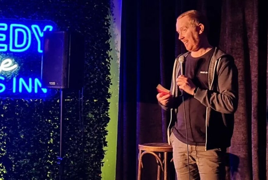Comedian holding a microphone on a small curtained stage, a neon sign partially visible to his right.