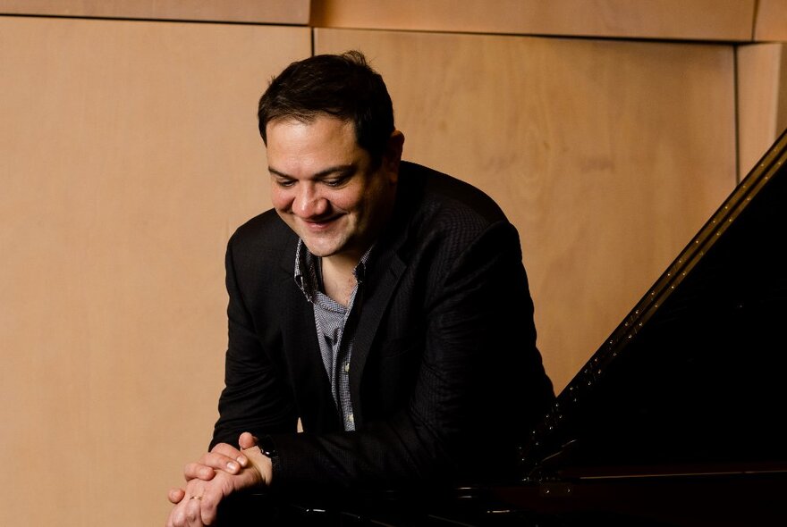 Pianist Coady Green, a man seated next to a piano, gazing downwards with a slight smile on his face, his hands clasped together and resting on his knee.