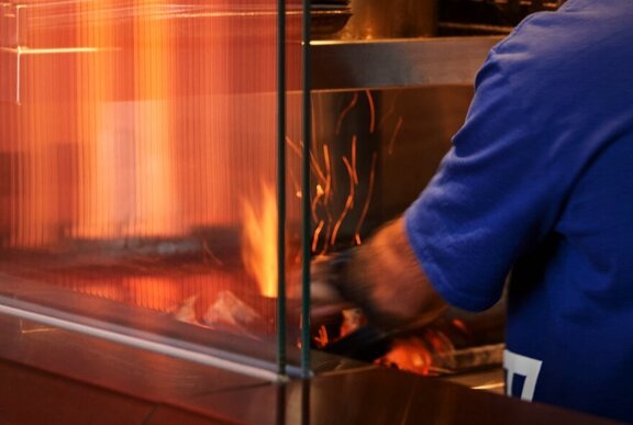 A chef in an open-concept kitchen at a restaurant with a fiery BBQ grill.