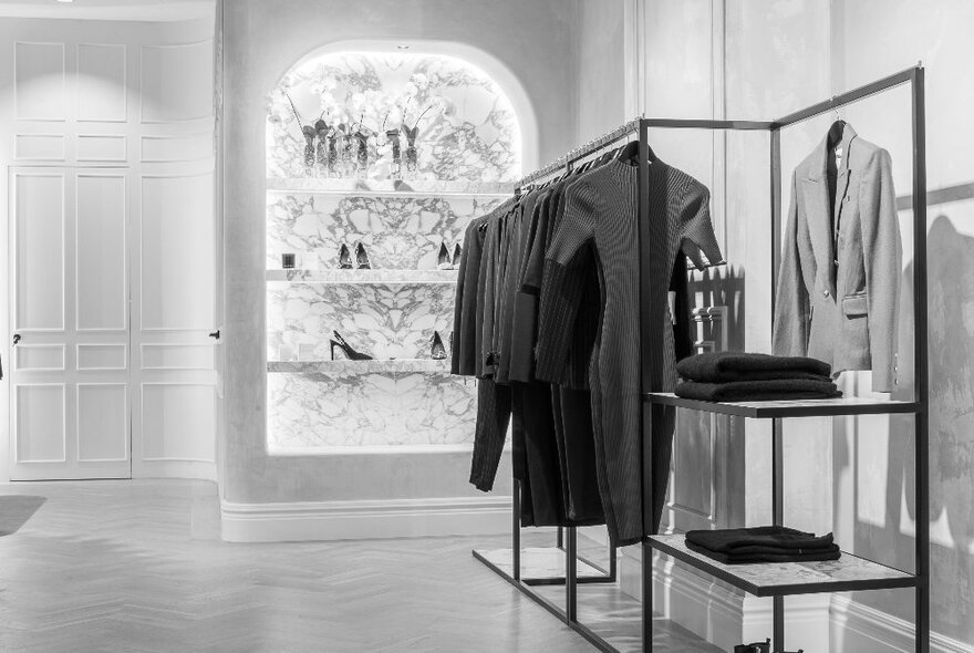 Camilla and Marc interior with sparse racks of clothing and a feature wall shelf with shoes in the background; black and white image.