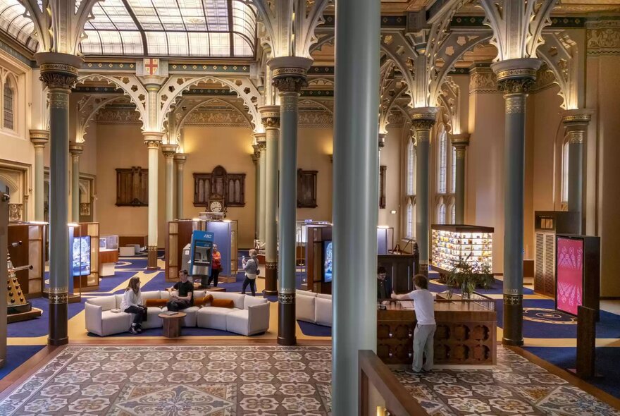 Interior of the grand, ornately designed main hall of the ANZ Banking Museum, featuring high ceilings supported by detailed columns and arches, decorative tiled flooring, display cases, and couches.