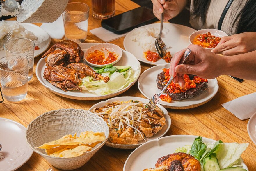 Plates of Indonesian food on a table with people serving themselves with forks.