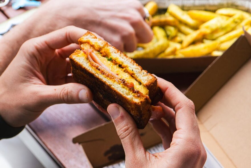 A person's hands holding a toasted sandwich with egg and bacon fillings, a cardboard box filled with chips behind it.