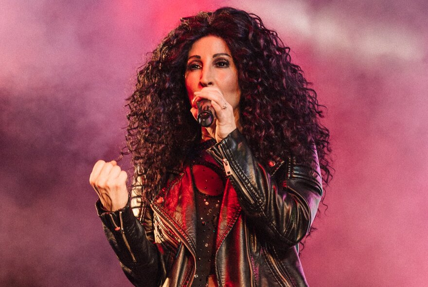 A tribute performer dressed as Cher singing soulfully with a red lit background.