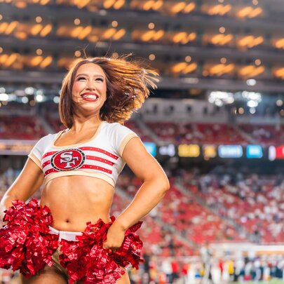 49ers cheerleader with brief top holding red streamers in front of large crowd in an arena.