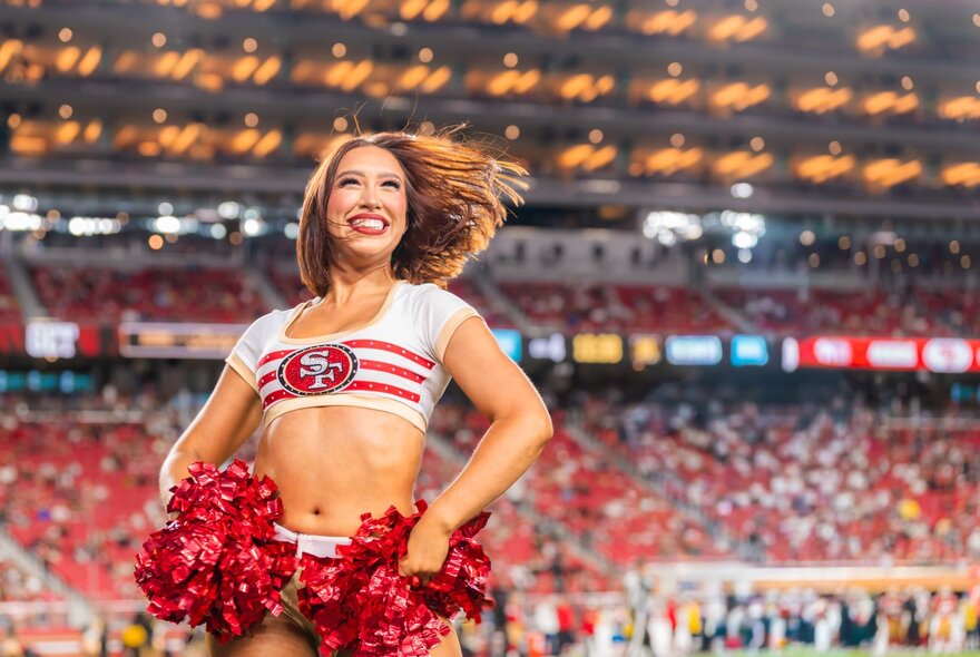 49ers cheerleader with brief top holding red streamers in front of large crowd in an arena.