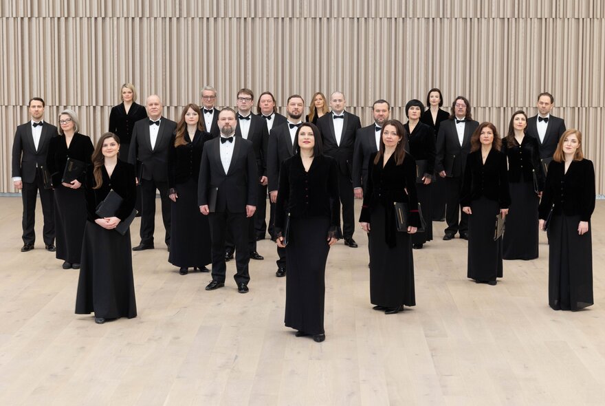Classical choir standing wearing black gowns and tuxedos in a beige space.