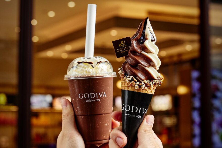 Hands holding up a soft serve chocolate ice-cream and a chocolate drink, both items in Godiva branded packaging.