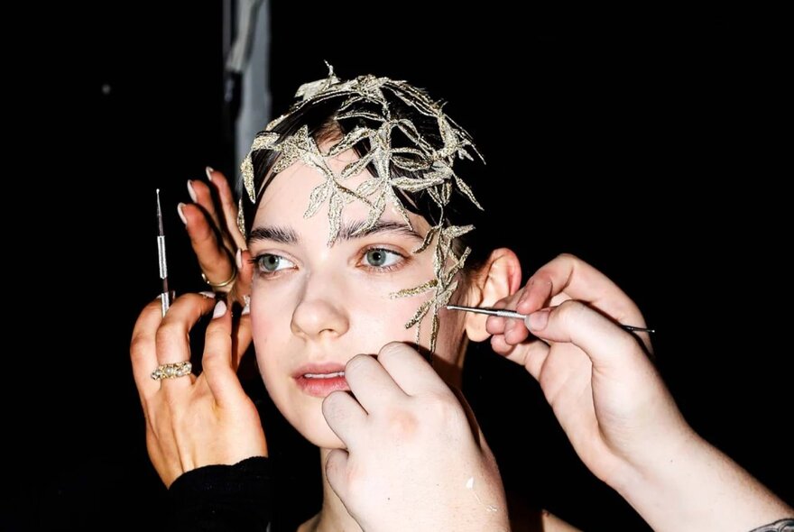 A model having makeup applied and her hair arranged under a head covering, by two sets of hands, as part of preparations for a fashion runway show.
