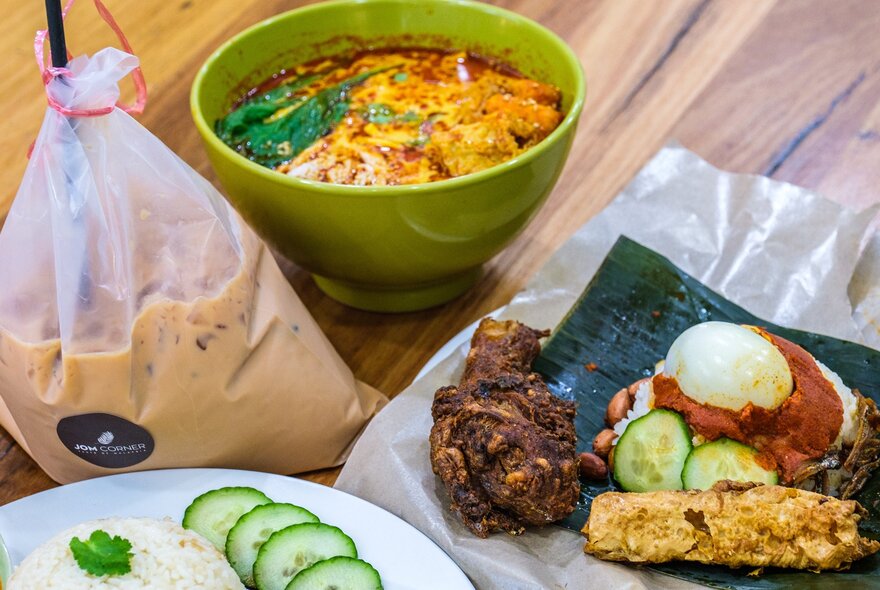 A bag of Malaysian iced tea, a bowl of laksa and a plate with nasi lemak featuring rice, egg, chicken, cucumber and sambal.
