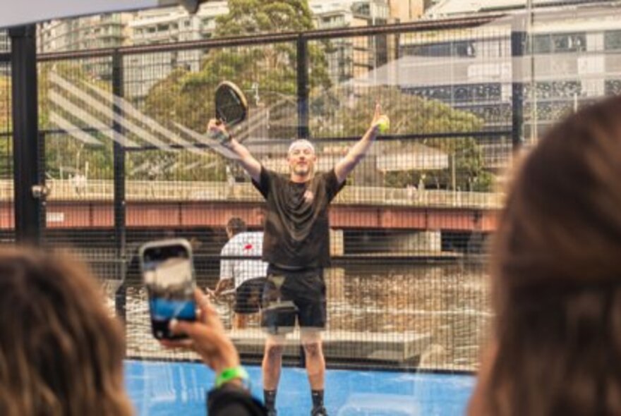 A man raising his arms in celebration while holding a racquet and ball on a blue enclosed court.