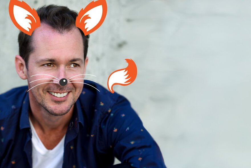 Actor and singer Mike McLeish wearing a blue shirt, looking sideways and with fox ears, fox whiskers and a fox tail hand drawn over his face and body.