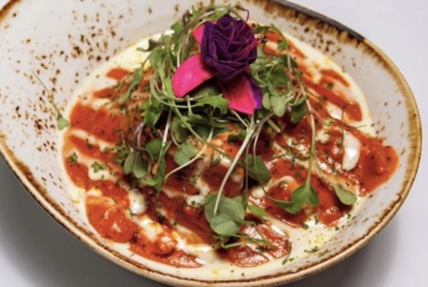 A Nepalese curry served in an earthenware dish with a white sauce drizzled on top; microgreens and a purple flower for garnish.