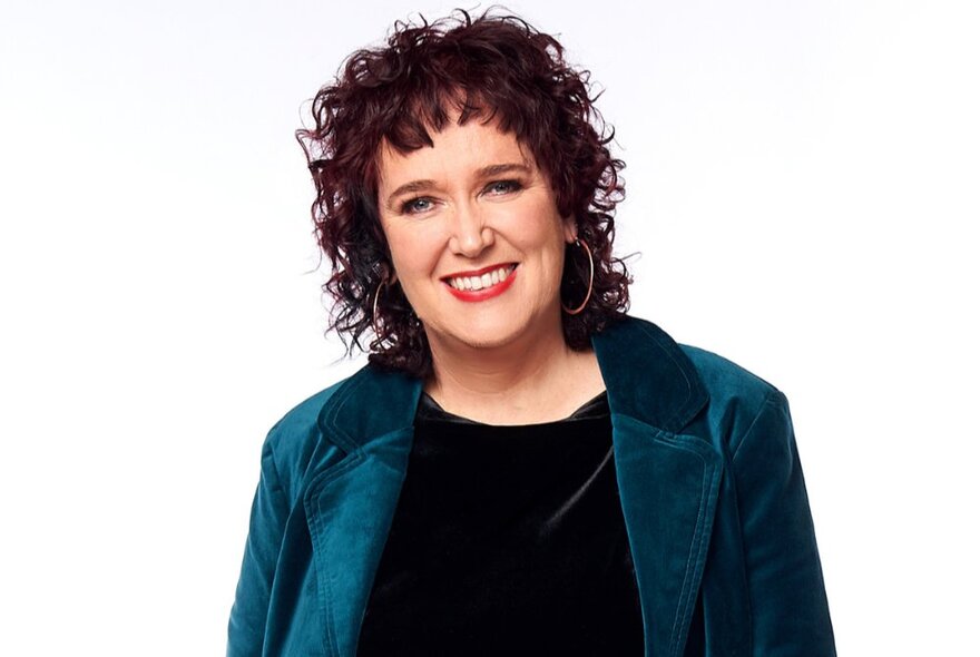 Journalist Natasha Mitchell, a smiling woman with curly brown hair, wearing large hoop earrings and a teal coloured velvet jacket.
