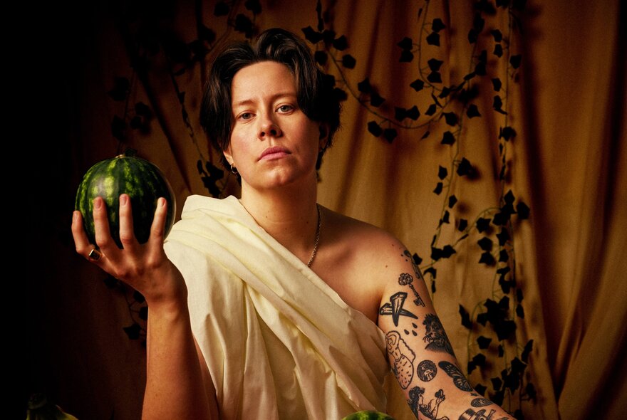 Chloe Petts, wearing a sheet wrapped around her like a toga, with a neutral expression, and holding a whole mini green watermelon in her upturned hand.