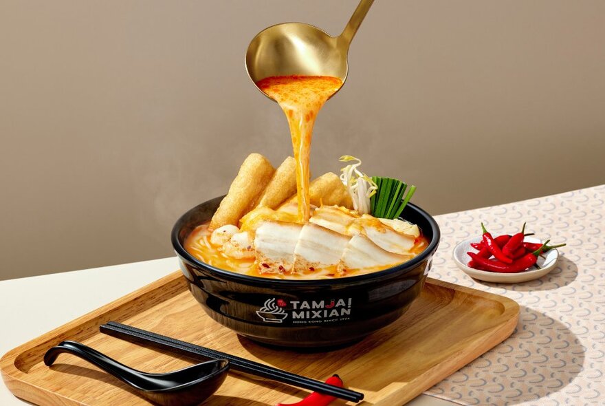 Someone pouring a final ladle into a bowl of noodle soup with condiments including small bowl of chillies.