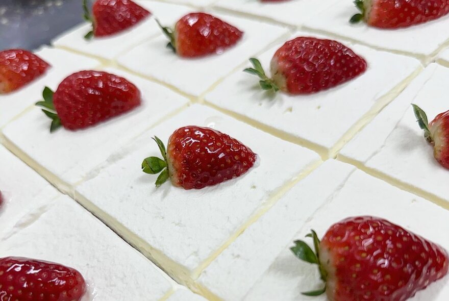 Squares of cream covered sweets all with a strawberry on top.