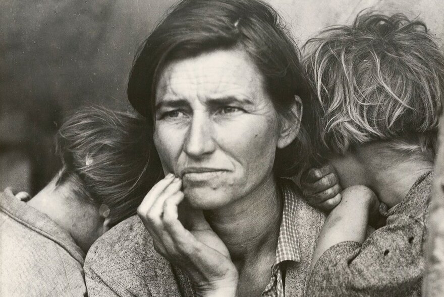 A black and white portrait photograph of a worried young mother with her two children hiding their faces while hugging her, circal 1940.