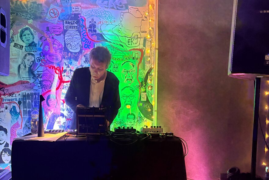 DJ at a mixing desk with psychedelic-coloured graffiti in the background.